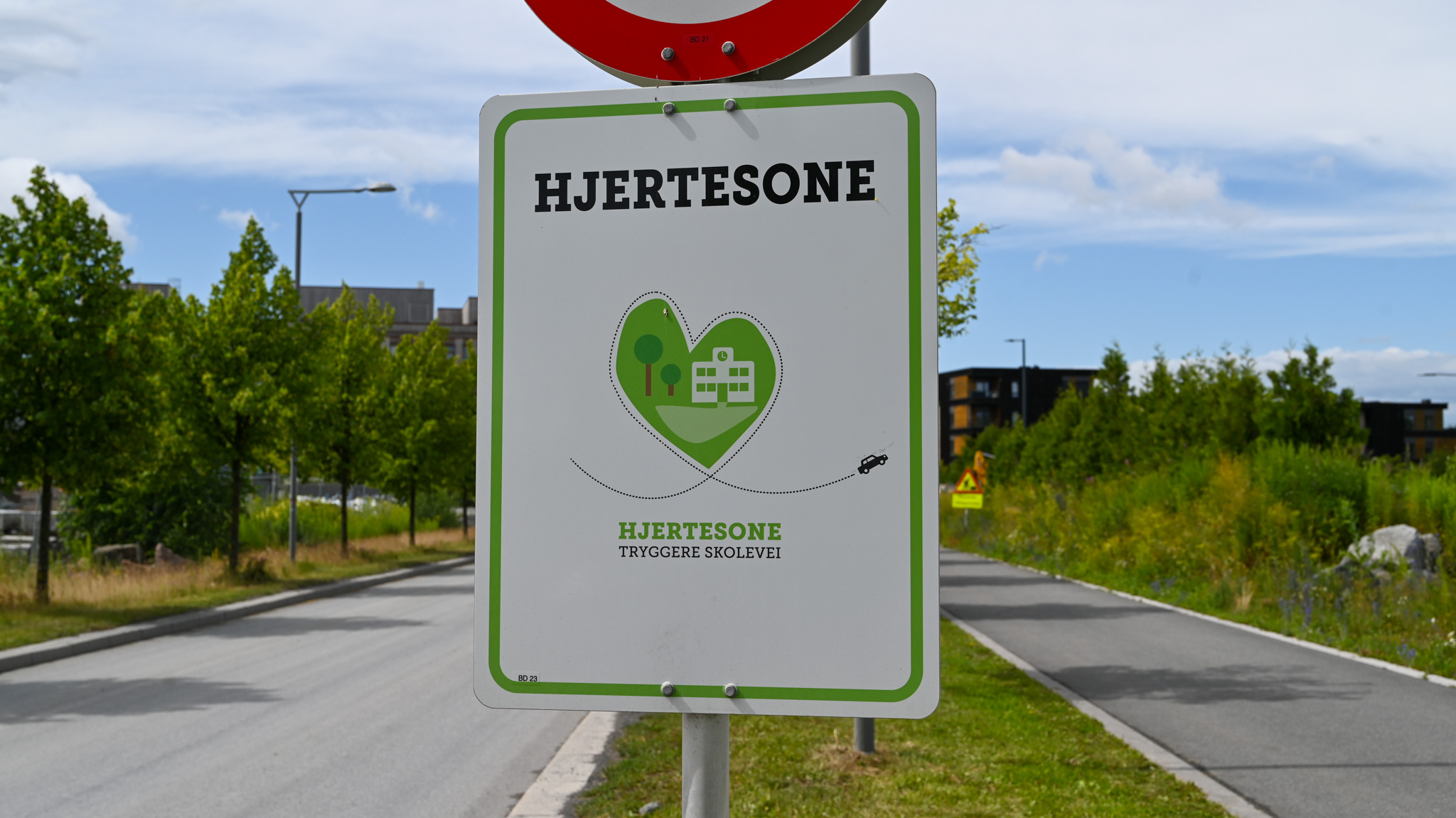 Et skilt med teksten 'HJERTESONE' og en illustrasjon av et grønt hjerte med trær og bygninger. Under hjertet står det 'HJERTESONE TRYGGERE SKOLEVEI'. Skiltet står ved en gate omgitt av trær og bygninger i bakgrunnen.
